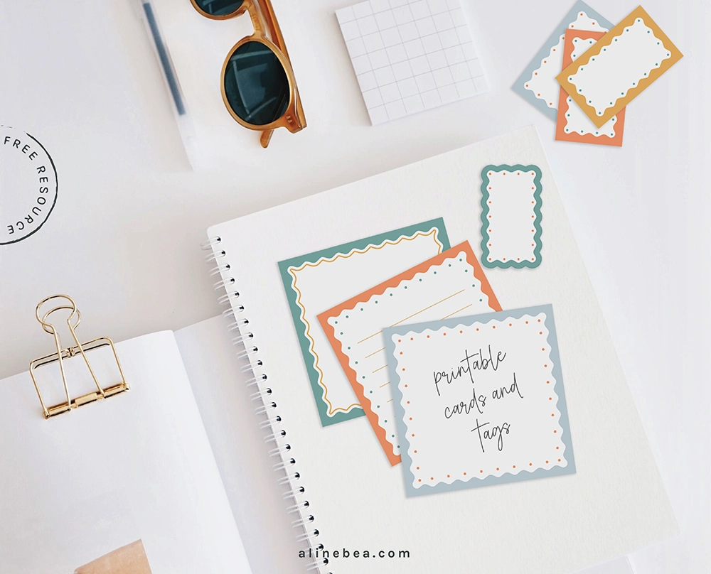 Notecards and tags with colorful abstract graphics around their borders, on top of a blank notebook, on a white table. On top of the table there is an open book and sun glasses.