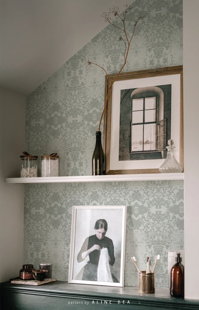 Small section of a low wooden cabinet with shelves above it, in the corner of a living room. The shelves contain a framed picture of a woman embroidering, personal objects and another frame with a picture of a window. The wall in the background has a wallpaper from Spark collection, designed by Aline Bea.