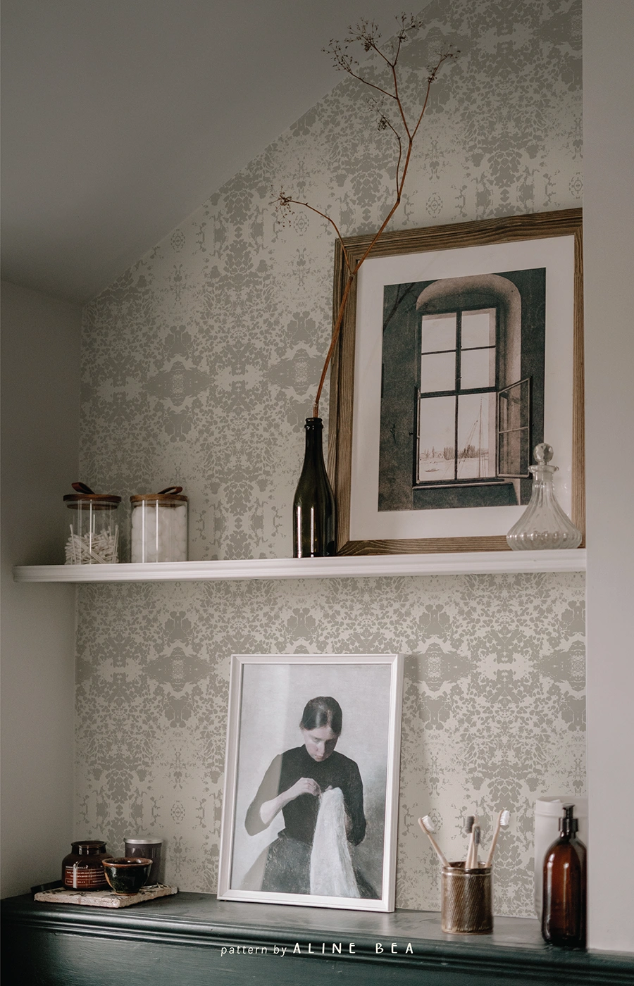 A section of a vintage cabinet on bottom, with a framed picture of a woman sewing and small decorative objects on top. A shelf in the middle of the picture, with a wood frame, a vase and more decorative objects on top. Everything is brought together by the wall with a damask-like wallpaper in the background.