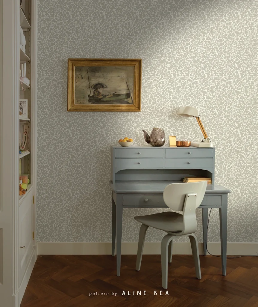 Vintage home-office scene, displaying a wall covered by a decorative wallpaper by Aline Bea, floor to celling cabinet with open shelves on the left, a vintage style wooden working table with small drawers and a lamp on top and a wooden chair in front of it. An antique hand painted art in a golden frame is hanging on the wall above the table.