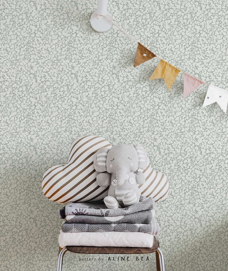 Nursery room scene, displaying a chair with comfy blankets folded on top of it, and a small stuffed elephant made of crochet on top of the blankets. Colored small fabric flags are hanging in the corner of the frame. The wall behind it all is covered by a decorative wallpaper by Aline Bea.