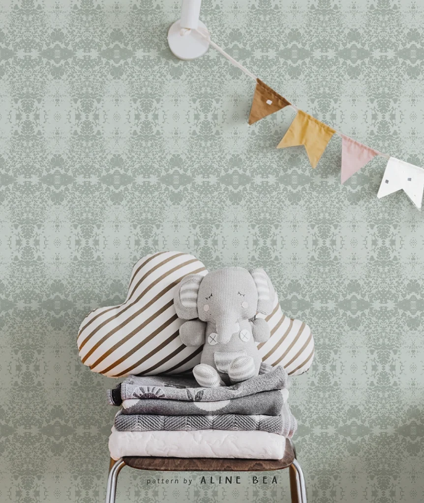 Nursery room scene with a wall covered by a delicate wallpaper design by Aline Bea, a chair in front of it, containing some folded blankets and a small stuffed elephant on top. Small fabric flags are hanging in a line in the corner of the image.