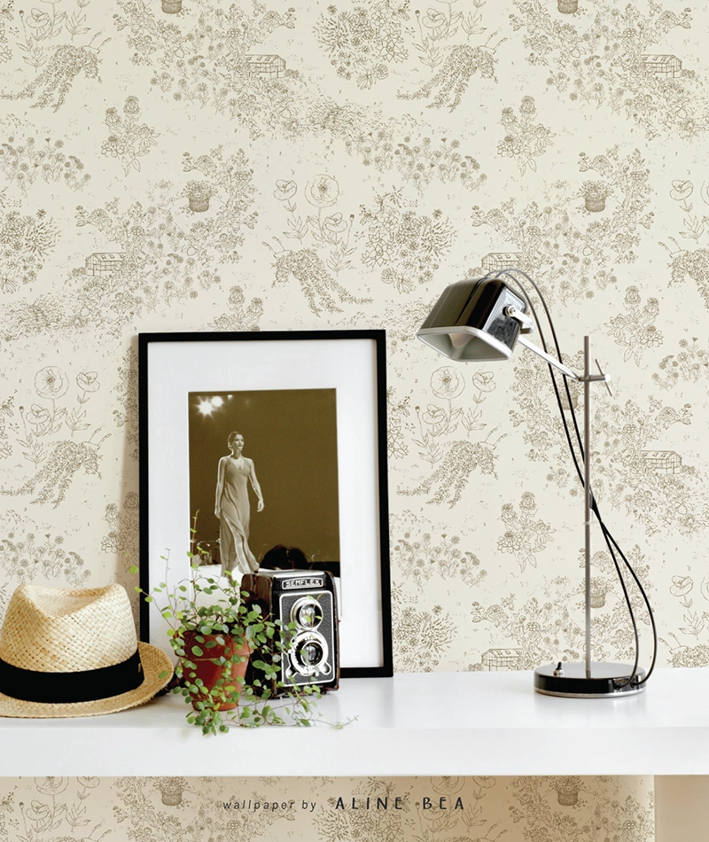 A section of a wall in a living room, displaying a wallpaper on toile de jouy style, designed by Aline Bea. On front of it there's a shelf, with a framed picture, an antique camera, a table lamp and a hat on it.