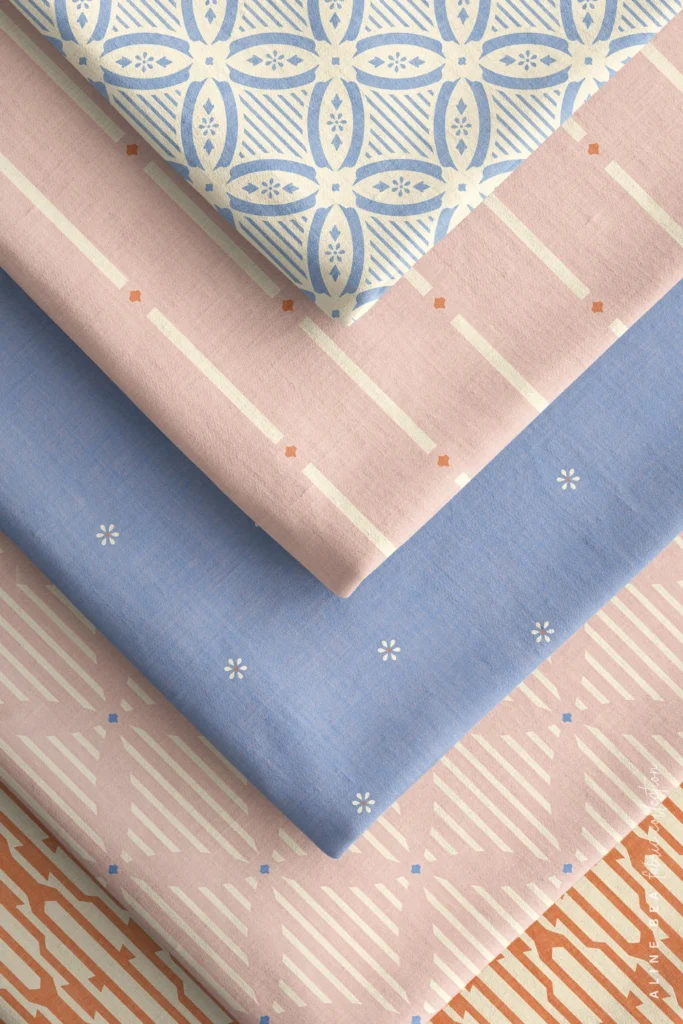 Five fabric swatches folded and stacked diagonally: a blue-and-cream quatrefoil lattice on top, then a pink and cream dashed stripes, a solid dusty blue fabric scattered with tiny white star-shaped florals, a pink fabric with cream striped lozenges, and an orange geometric trellis visible at the bottom edge.